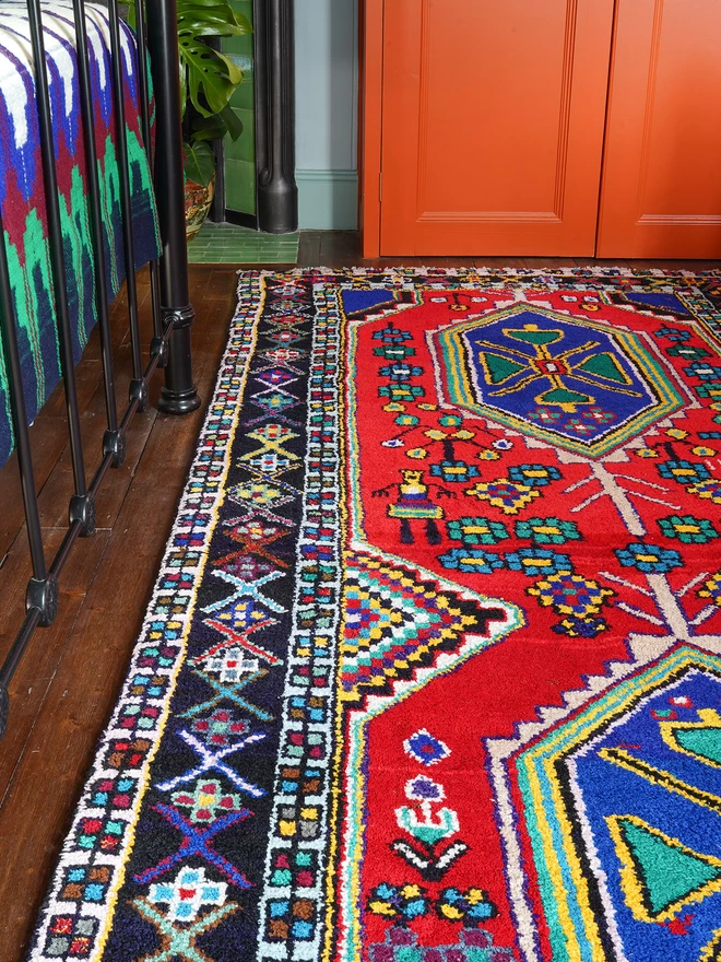 Brightly coloured Persian vintage rug, in a red, green and blue pattern in a bedroom with orange wardrobe,  and iron bed