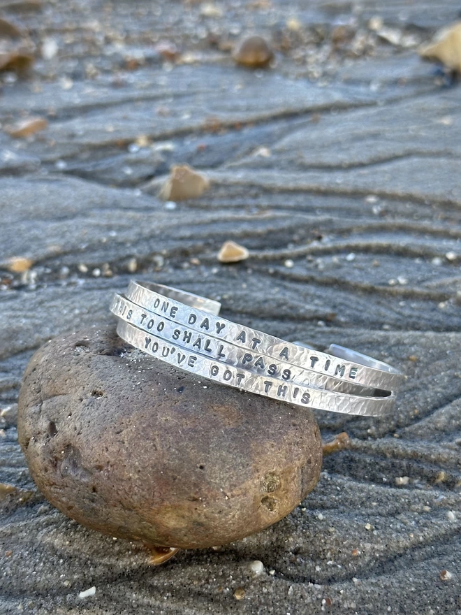 A stack of three sterling silver hammered cuff bangles with stamped text, resting on a rock that is placed on the sand of a beach with ripples from the water visible around it.
