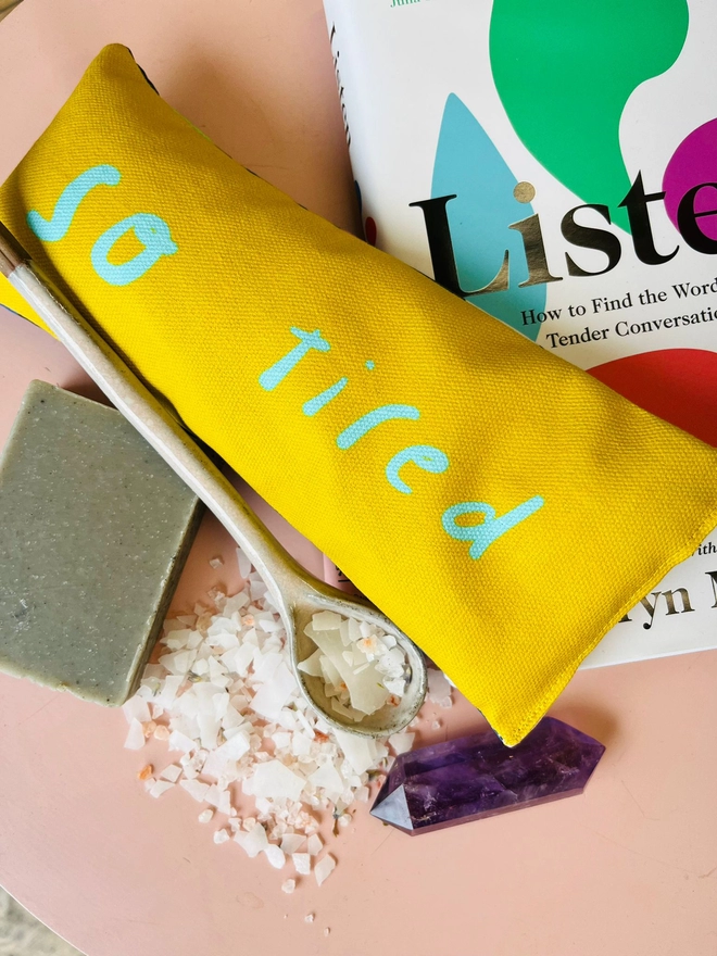 Flatlay of relaxing items including lavender eye pillow which says 'so tired' - yellow fabric with neon turquoise letters. Crystals, bath salts, and book surround it. 
