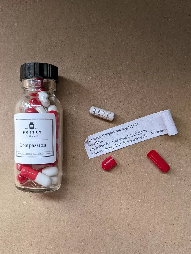 a glass bottle of red and white Compassion poetry pills on brown paper, with an open pill next to it and an unrolled poetry quote