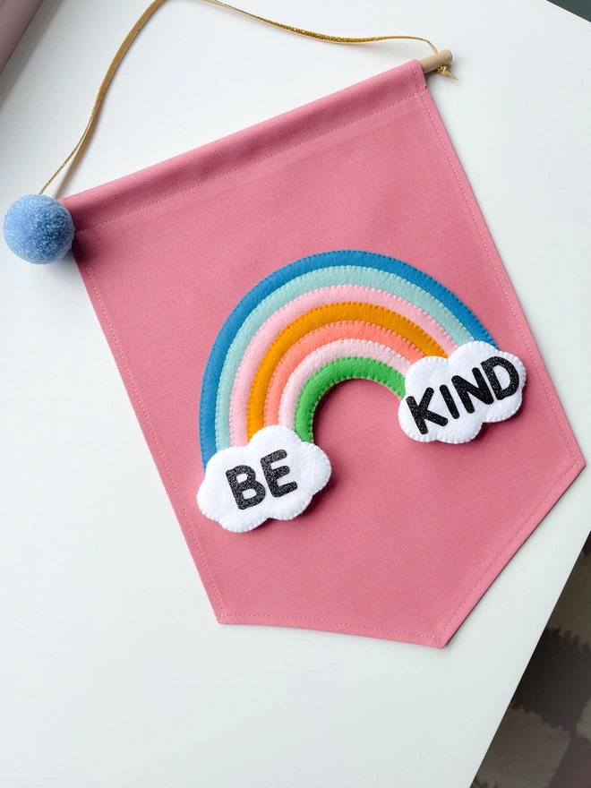 Pink banner with a rainbow and two clouds saying the words be kind