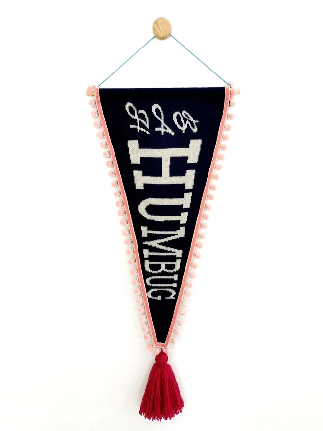 A product image of a navy knitted pennant flag with the words ‘Bah Humbug’ written in a white sparkly retro text down it. The banner is trimmed with blush pink pom pom trim and has an oversize raspberry pink tassel hanging from the bottom of it. The whole banner is suspended from a wooden wall dot by a bright teal nylon cord.