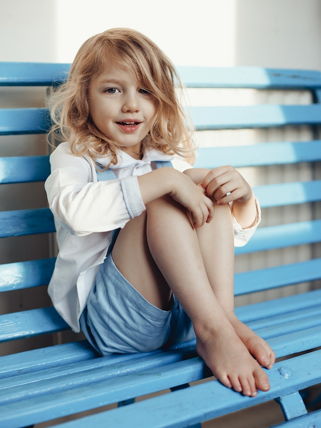 A little boy sits on a blue bench wearing a white peter pan  collared shirt and blue shorts with braces