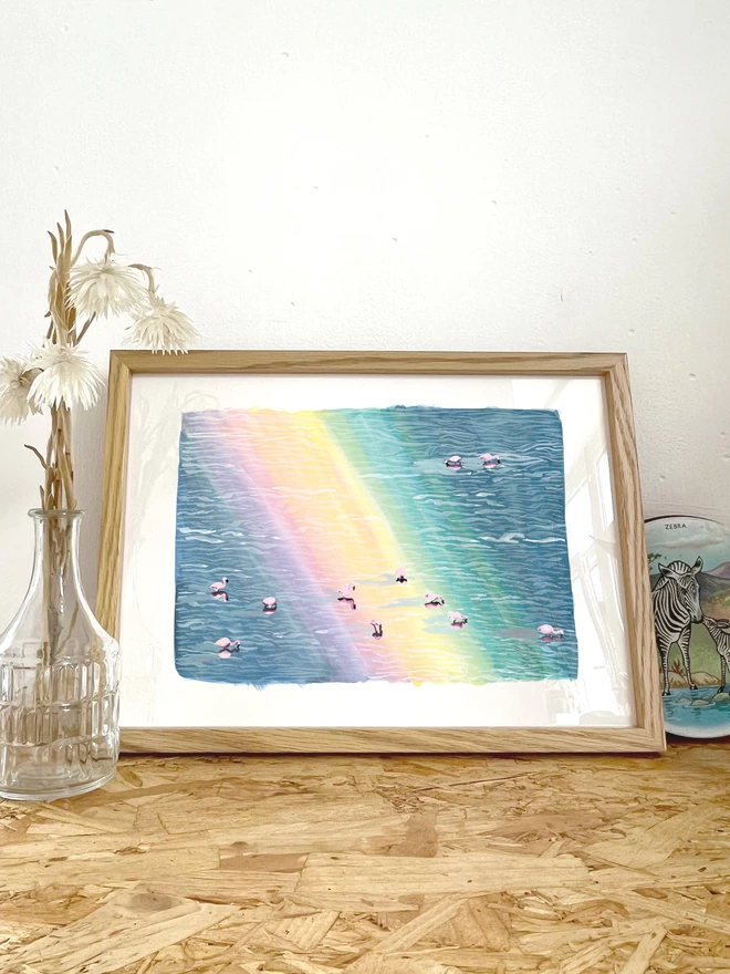 a print featuring a colourful illustration of flamingos in a lake with a rainbow on the water in a frame next to some flowers and a tin with a zebra on it