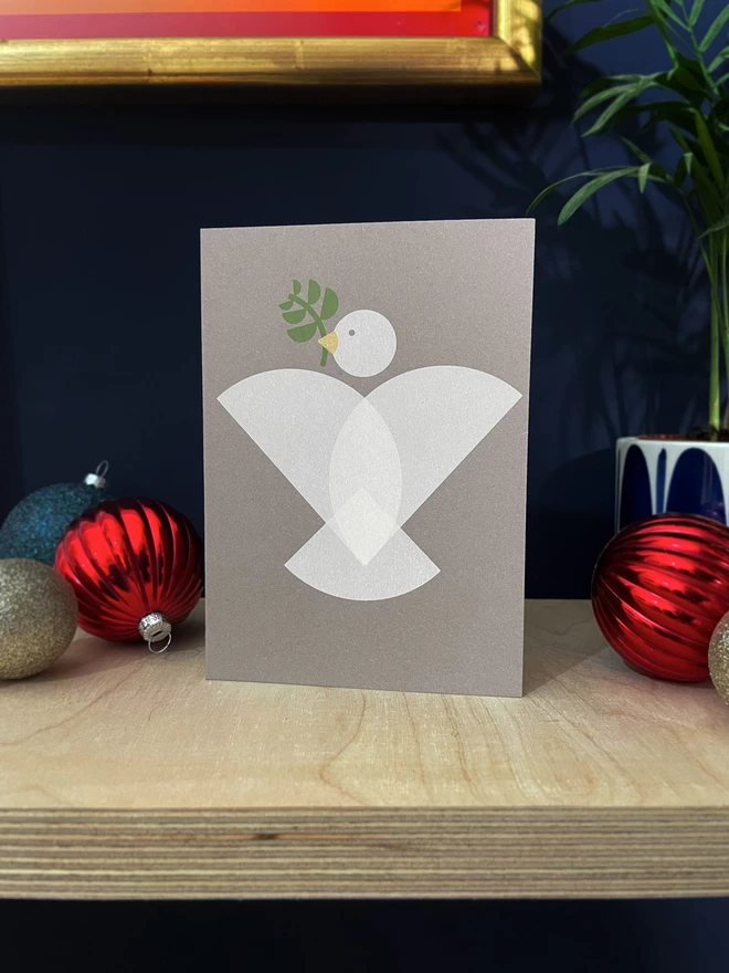  Geometric shapes make up a dove design on this handprinted christmas card, with an olive branch and gold beak. Stood on a plywood shelf with baubles, foliage and a hint of a golden frame  around.