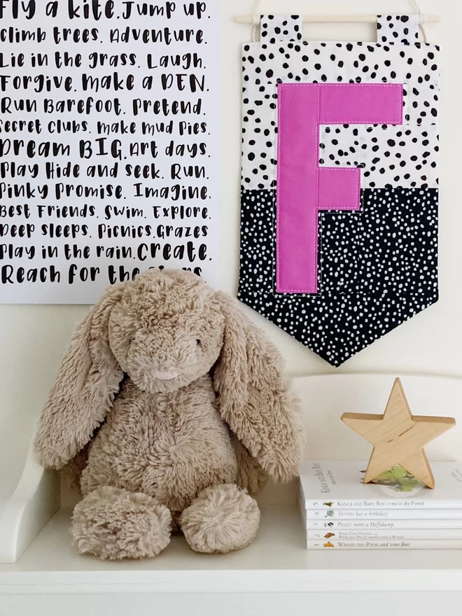 Cooper and Fred colour block quilted wall hanging with spotty background and a pink letter 'F', hung on a wall above a shelf with decorative items.