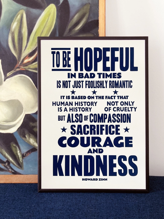Framed typographic print of a quote by Howard Zinn. It reads "To be hopeful in bad times is not just foolishly romantic, it is based on the fact that human history is a history not only of cruelty but also of compassion, sacrifice, courage and kindness". Navy blue text on white paper. The print rests against a painting of white flowers on a dark blue background.