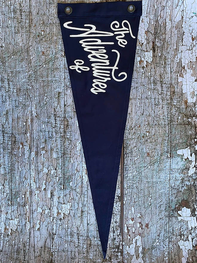 A navy coloured pennant flag hung vertically on a distressed wooden wall. In ivory lettering are the words ‘The Adventures of'