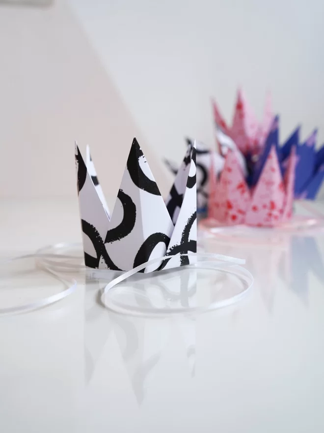 A selection of paper party crowns in various patterns and colours on a reflective glass surface.