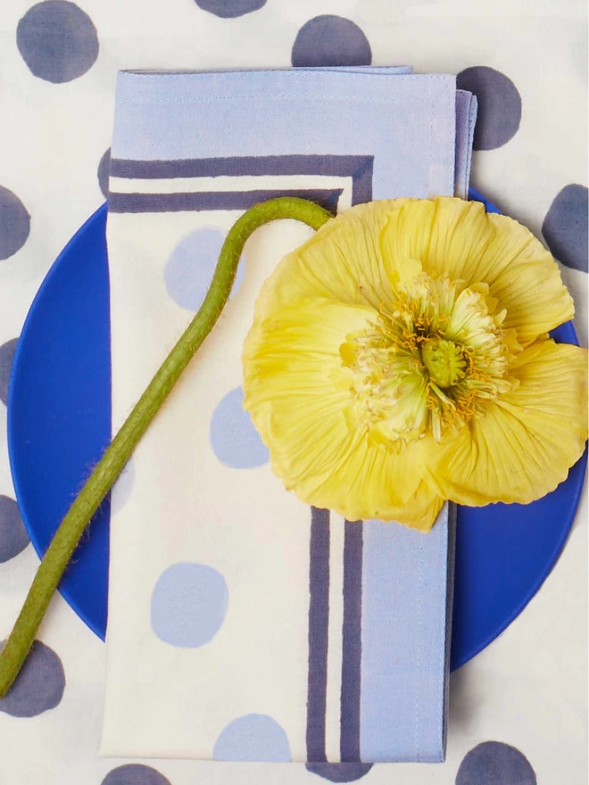 Single pale blue block printed napkin with an irregular dot pattern design, on a bright blue plate and with a yellow flower resting on top