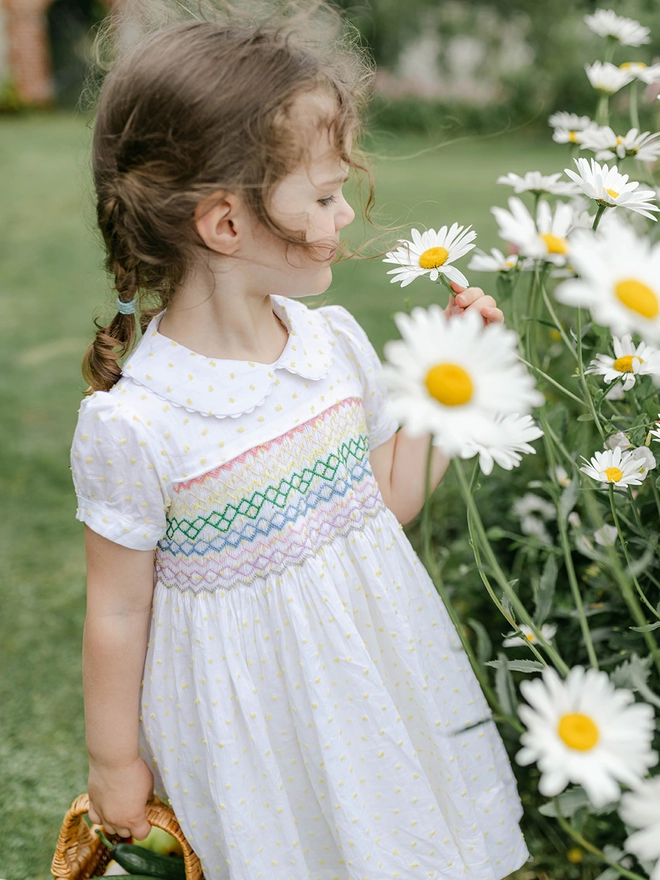 A girl looks at flowers in a white and yellow swiss dot dress with a peter pan collar and rainbow smocking