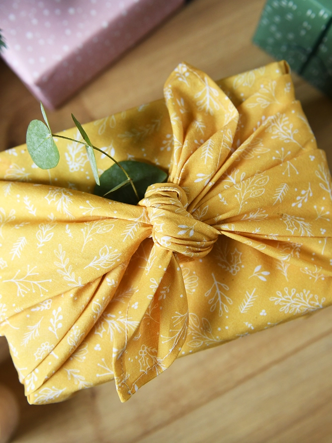 A gift wrapped in yellow cotton fabric wrap with a botanical design, and a small posy of greenery tucked into the knot, stands on a wooden table.