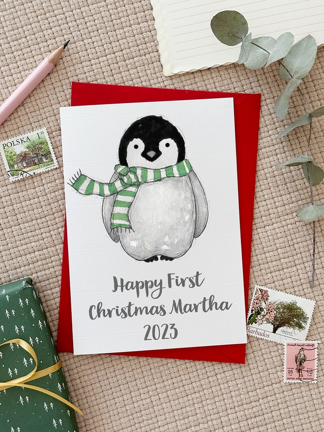 A first Christmas greetings card with an illustrated penguin design lays on a red envelope beside various stationery items.