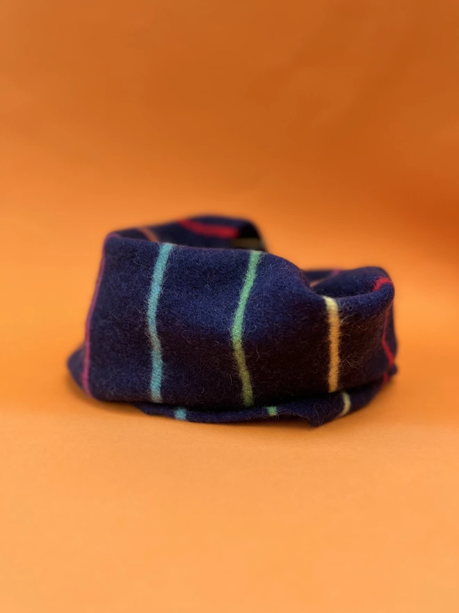 Navy knitted snood with colourful thin stripes shown from side angle on orange background