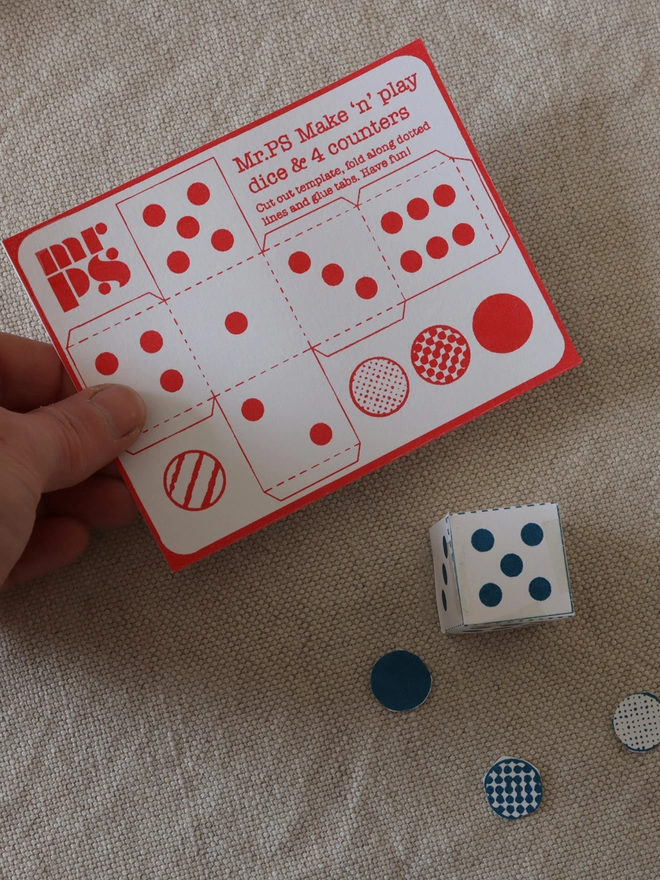 A hand holding the die and counters card which comes with the Mr.PS Snakes & Ladders hankie
