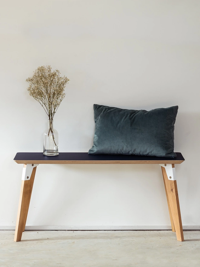 A stylish and minimalist bench with black Fenix top, white coloured steel brackets and solid oak legs with a cushion and vase on top