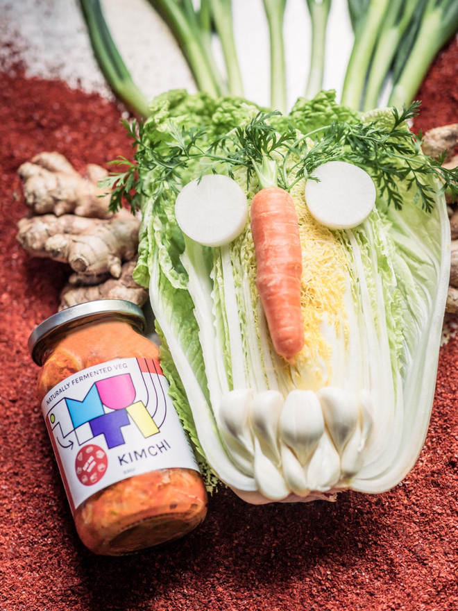 A Jar Of Muti Kimchi On A Bed Of Chilli Flakes Beside A Jungle Mask Made Of The Fresh Ingredients Used In Muti Kimchi.