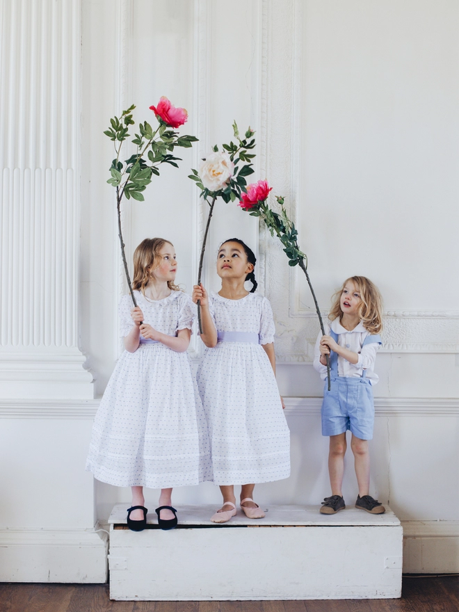 A little boy wearing a white peter pan  collared shirt and blue shorts with braces stands on a box holding a flower alongside two girls wearing white dresses with blue sashes who are also holding flowers