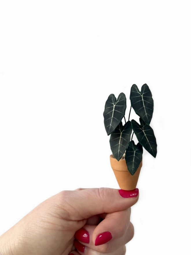 A miniature replica Alocasia Frydek ornament made from paper in a terracotta pot balanced on a thumbnail