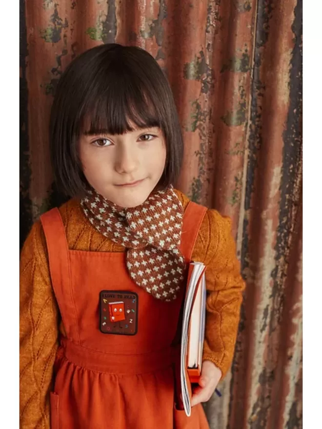 A kid holding some books wears an orange pinafore with the I Love To Read patch.