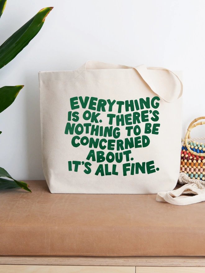 a large natural canvas tote bag bearing the words Everything is OK. There's nothing to be concerned about. It's all fine stood up on a bench