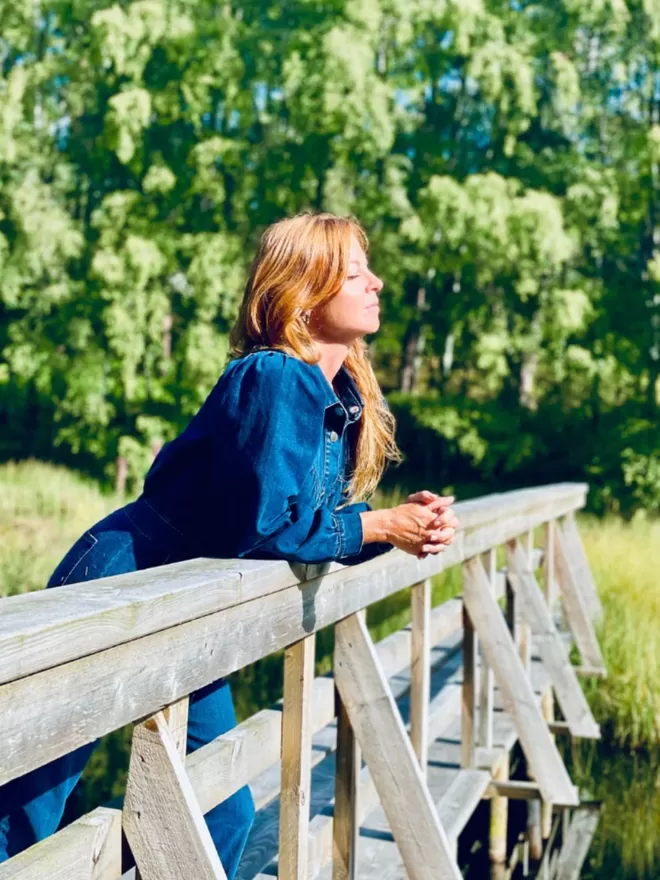 The Maggie Denim Jumpsuit worn by the model outside on a bridge over water in the sun