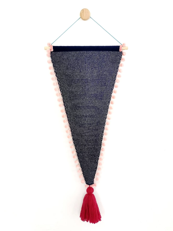 A product image showing the reverse of a navy knitted pennant flag. The back of the banner is knitted into a navy and white birdseye check and is trimmed with blush pink velvet pom pom trim. The point of the banner has an oversize raspberry pink tassel hanging from the bottom and the whole flag  is suspended from a wooden wall dot by a bright teal nylon cord.