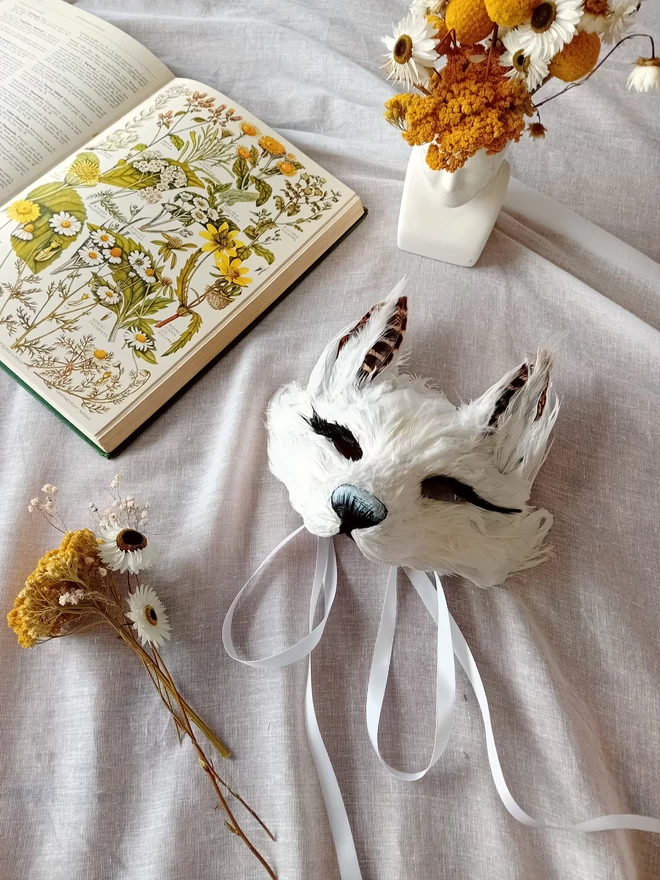 A kid's luxury white fox mask flat lay with yellow and white flowers and a botanical book