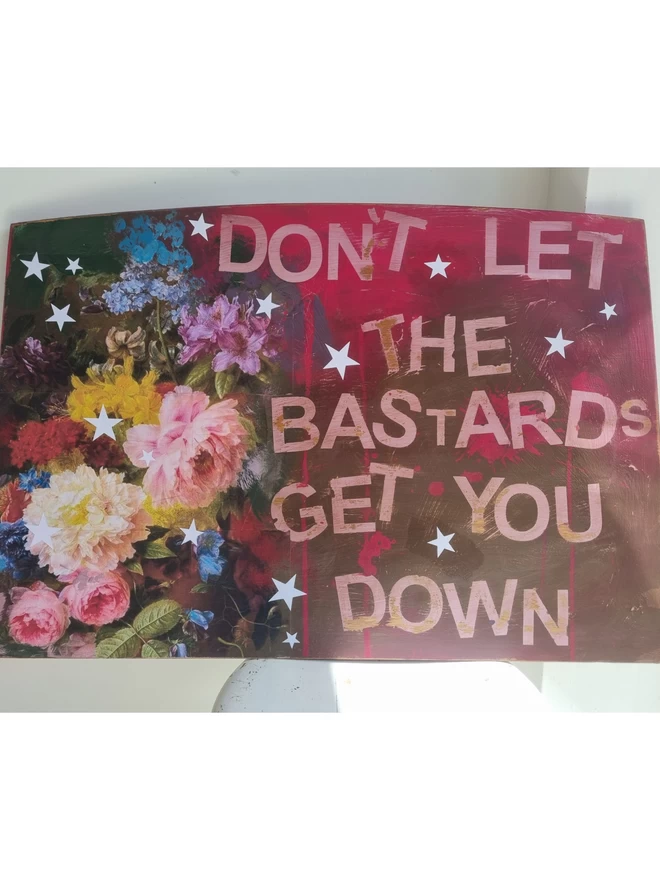 Art panel made from an upcycled table top and decorated using decoupage florals in pinks and browns. The text reads ‘don’t let the bastards get you down’ in the hot pink undercoat. Decorated with stars.