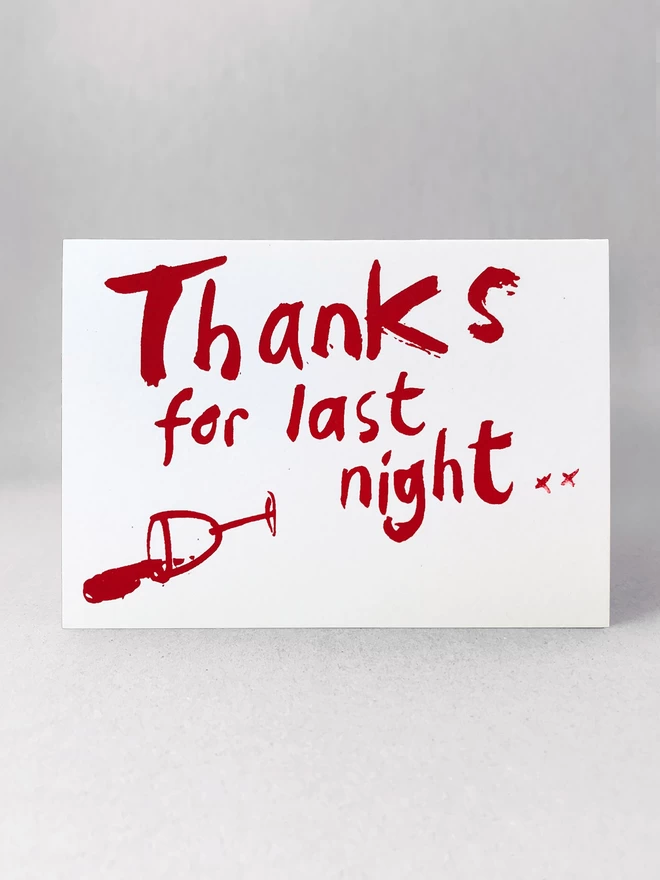 Thanks for Last night in a handwritten scrawl is screenprinted across this greetings card in claret red, with a knocked over wine glass besides. The card is stood  in a light grey studio background, flat on,