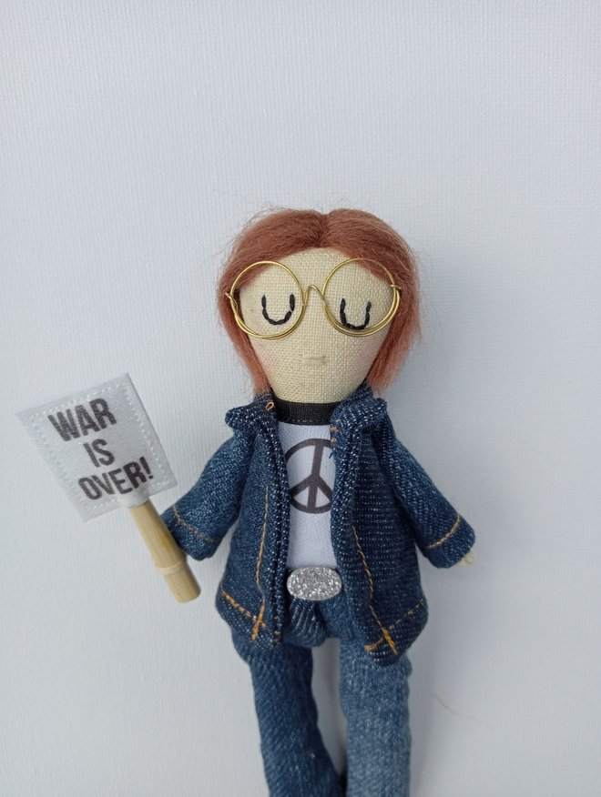 John Lennon mini decorative icon doll against a white background John is dressed in denim flares and denim over shirt with white tee emblazoned with black peace sign in his right hand is a white placard with black text reading war is over John has chin length shaggy hair and gold wire framed circular glasses 