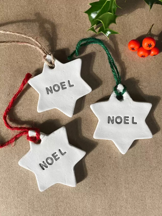 3 star shaped clay tags with 'NOEL' stamped in the centre of the star. Each finished with either red, brown or green twine. Brown paper parcel background with a holly and berries in the top half of the image.