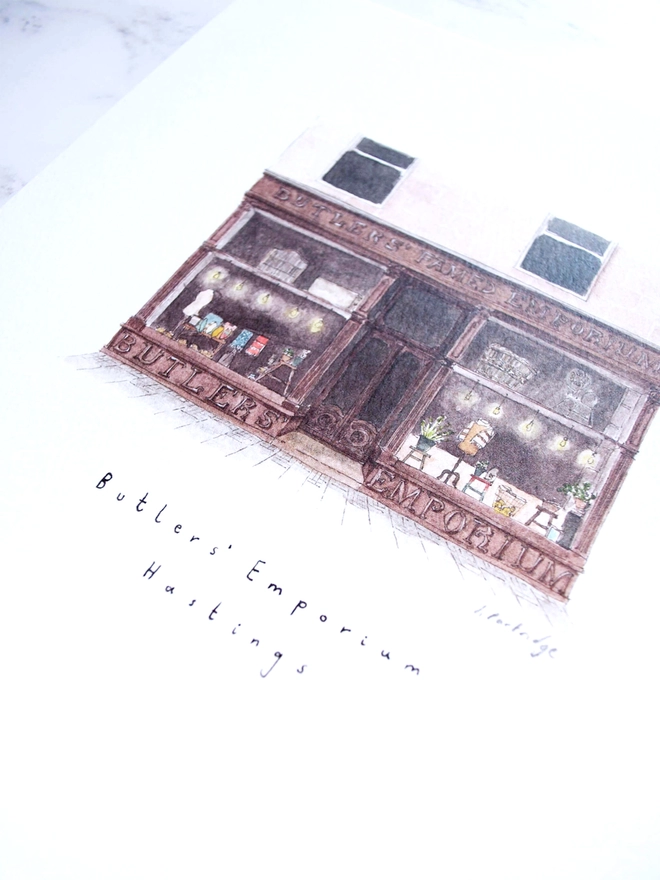 Watercolour illustration of Butler’s Emporium shop in Hastings, a brown shopfront with beautiful big windows full of treasures, the shop sells homewares and gifts carefully curated from small independent and local businesses. The painting is on a white piece of paper with the name written below. The photo is at an angle showing some of the intricate detail of the painting. 