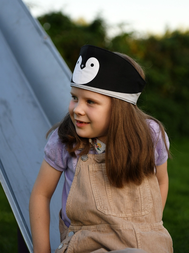 A young girl wearing beige dungarees and a handmade penguin fabric crown sits on a park slide.