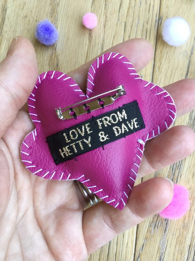 A hand holding a hot pink heart brooch, showing the reverse side with Love From Hetty & Dave label and brooch back