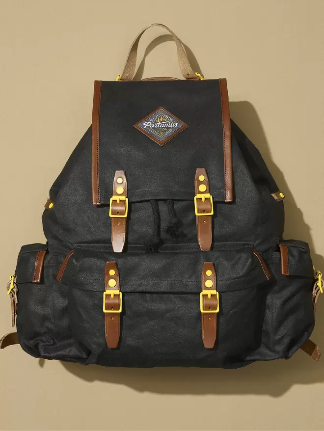 Black wax cotton Black Marten Rockness packpack with brown leather trim and yellow hardware on a plain taupe background.