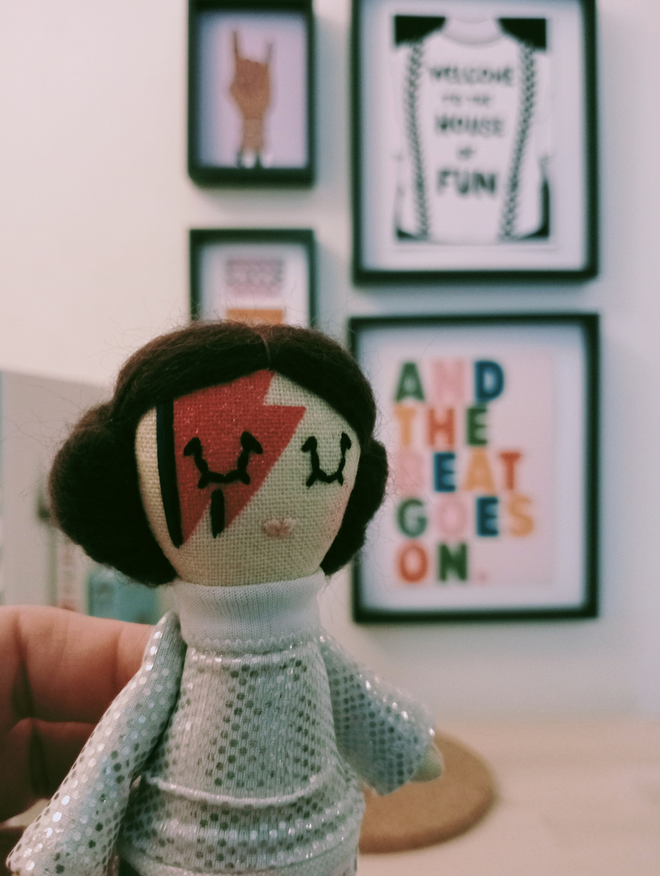Princess Leia mini doll with Ziggy stardust style make up held up to camera in front of a gallery wall of music themed prints