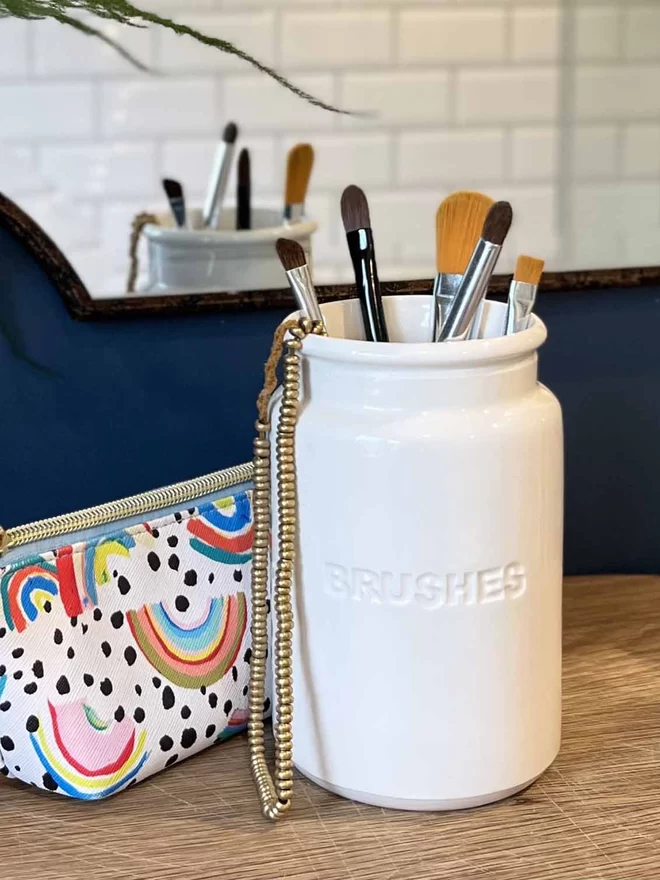 A handmade ceramic ‘brushes’ jar is in front of a mirror being used to hold makeup brushes.