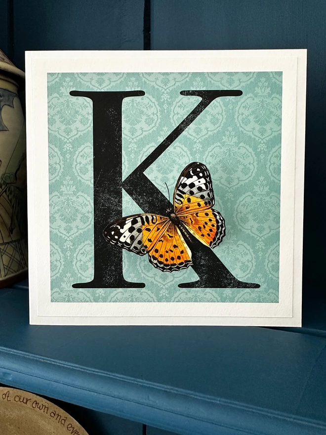 personalised birthday card with large letter on a blue coloured patterned background with a 3D paper cut butterfly. Card displayed on a blue shelf in home