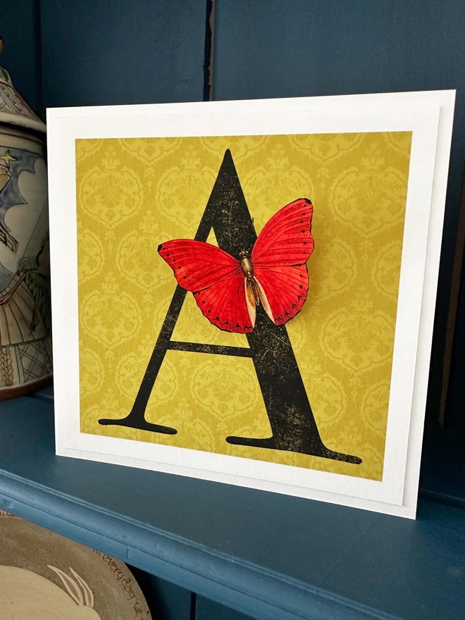 personalised birthday card with large letter on a gold coloured patterned background with a 3D paper cut butterfly. Card displayed on a blue shelf in home