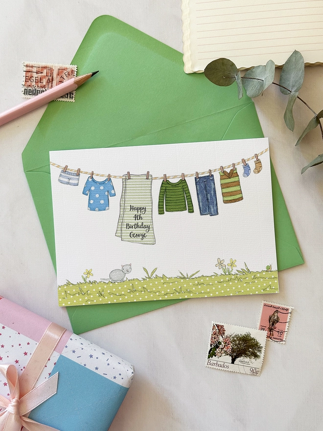 A personalised birthday greetings card with an illustrated washing line of children's clothes lays on a green envelope on a white desk.