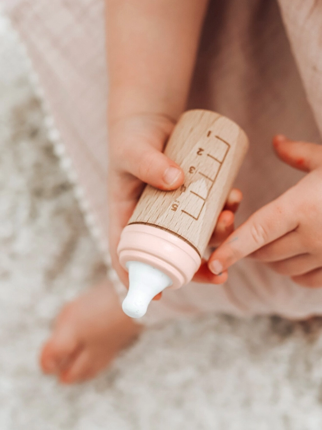 Wooden toy bottle in peach with measuring chart held by baby hands