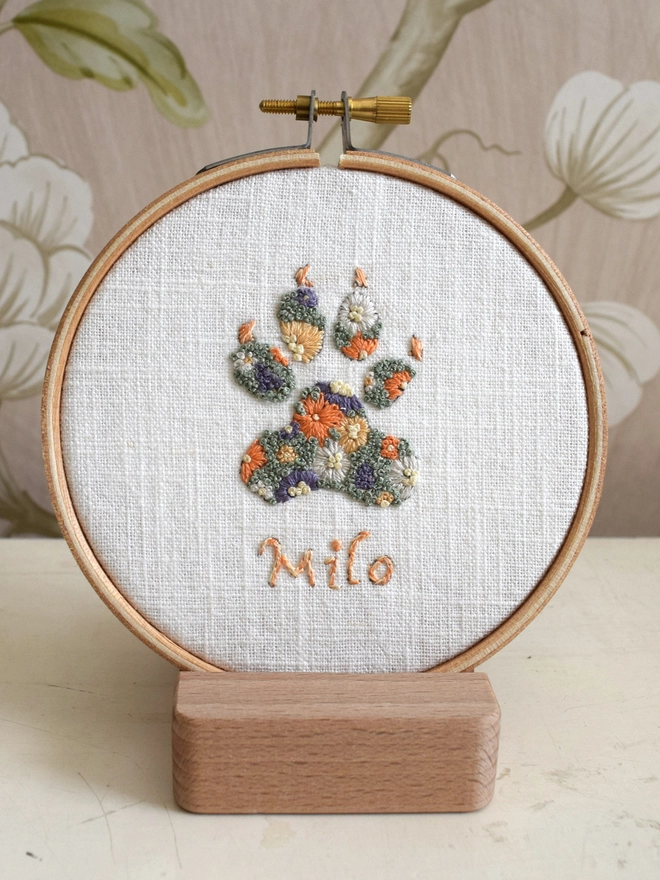 Sunshine Garden Dog Paw of Golden Yellows and Bright Orange Blossoms with Green French Knot grass background.  Displayed in an embroidery hoop on a wooden stand. 