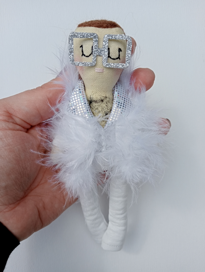 Elton John mini decorative icon doll held in a left hand for scale Elton is wearing a 1973 era inspired all white faux leather outfit trimmed with feathers and faux pearl buttons his glasses are silver glitter oversized square framed 