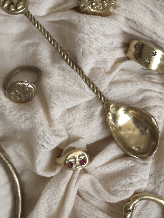 The image is of a collection of gold toned jewellery and a gold toned brass spoon laid out on a cream linen type fabric 