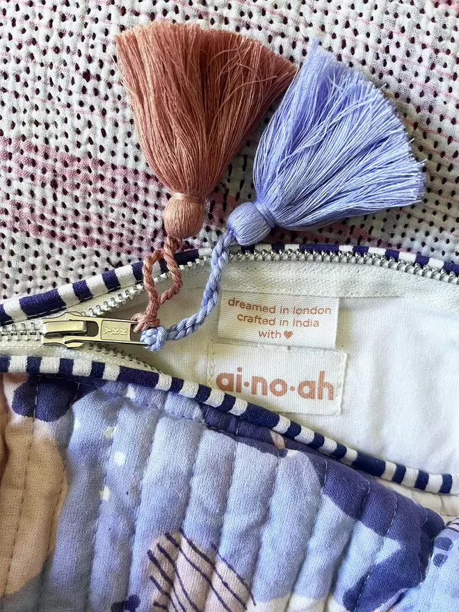 Close up of ainoah label inside a washbag , showing siukver zip and two tassels, one soft blue and one dusty pink