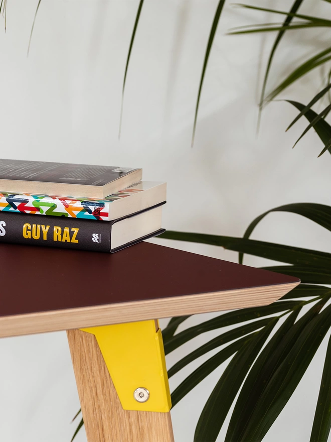 corner view of a stylish and minimalist standing height desk with burgundy Fenix top, yellow coloured steel brackets and solid oak legs, with a pile of books stacked up.