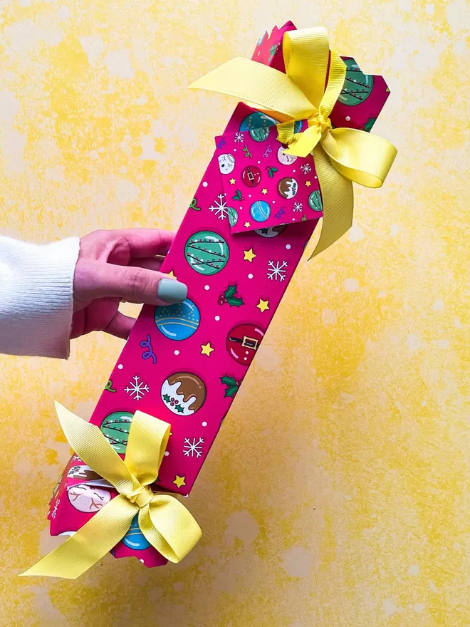 a person holding a red and yellow wrapped christmas cracker gift with cure illustrated macarons on a yellow background