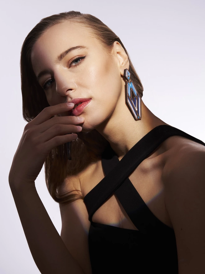 A woman modeling a hexagon shaped earrings on her left ear. Her hand is on the mouth. She's wearing a black top with a her hair sleek back.