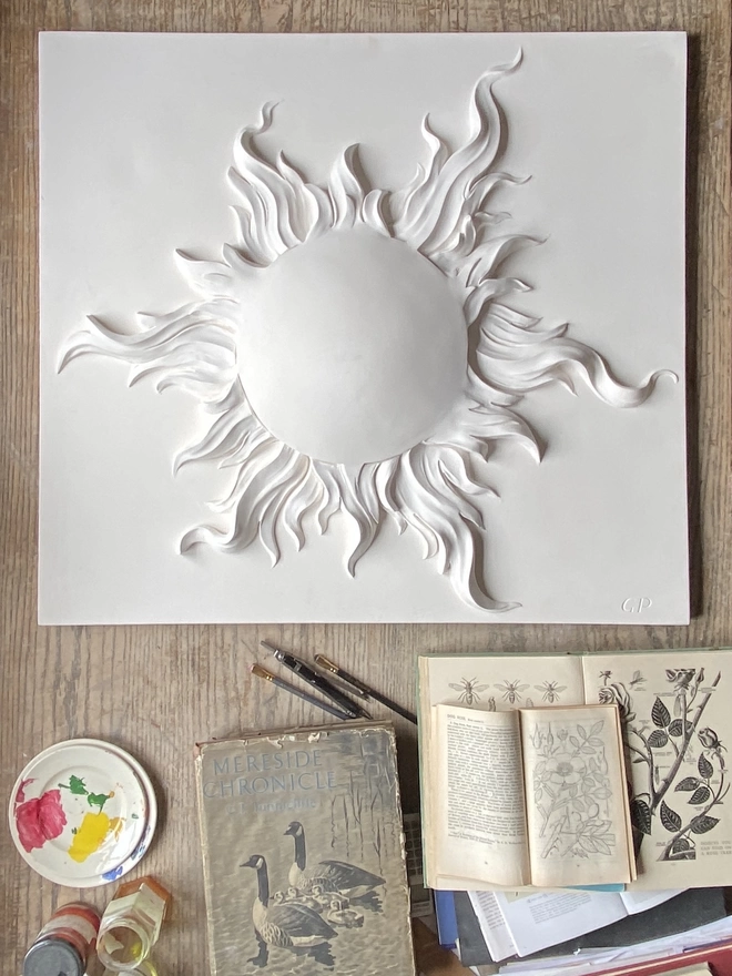Plaster bas-relief wall plaque of a sun on a table with books and art equipment underneath
